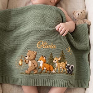 Könnte beinhalten: Eine salbeigrüne Strickdecke für Babys mit dem eingestickten Namen Olivia in goldener Schreibschrift. Das Motiv zeigt einen braunen Bären mit Laterne, einen Fuchs, ein Reh und ein Stinktier, die an Tannen unter einem kleinen Mond vorbeigehen.