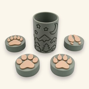 Puede incluir: Un recipiente cilíndrico gris con un diseño en relieve de montañas, árboles, estrellas y una luna creciente, acompañado de cuatro sellos circulares grises con huellas de patas y pezuñas de color rosa claro en sus superficies.