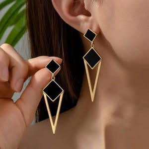 Puede incluir: Pendientes geométricos dorados con detalles cuadrados y de diamante negros. Presentan un pequeño cuadrado en la parte superior, un cuadrado negro más grande y un diseño dorado en forma de V. Un accesorio moderno y elegante.