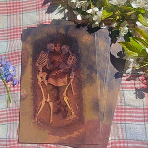 May include: A stack of art prints featuring a person embraced by two skeletons. The artwork has a warm color palette with a dark background. The prints are on a red and white checkered surface, with white flowers and green leaves in the background.