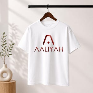 May include: A white short-sleeved t-shirt featuring a dark red graphic logo on the chest. The logo consists of an arched shape with a central dot above the text AALIYAH in a bold, sans-serif font. The shirt is displayed on a wooden hanger.