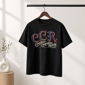 May include: A black short-sleeve t-shirt featuring a distressed graphic print on the chest. The design displays the letters CCR in large blue and red font, with the text Creedence Clearwater Revival written underneath in a smaller white script.
