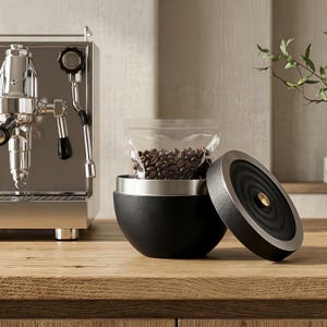 May include: A black and silver coffee bean storage container with a clear bag of coffee beans inside. The container is open, with the lid resting beside it. A stainless steel espresso machine is in the background, on a wooden surface.