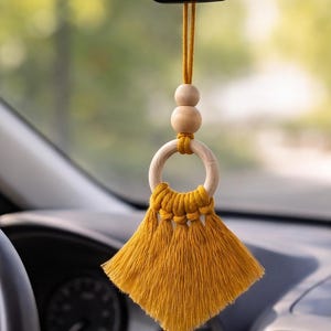 Può includere: Un accessorio per auto in macramè color giallo senape appeso a uno specchietto retrovisore. L'oggetto presenta due perline di legno chiare, lisce e rotonde sopra un anello di legno, con una nappa a ventaglio in corda di cotone giallo senape.
