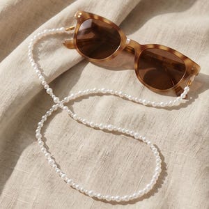 Pode incluir: Óculos de sol castanhos com armação tartaruga e uma corrente de pérolas brancas para óculos. Os óculos têm lentes castanhas e uma forma clássica. A corrente é feita de pequenas pérolas brancas redondas.