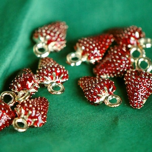 Pode incluir: Uma cole&ccedil;&atilde;o de pingentes em forma de morango vermelho e dourado com acabamento brilhante. Os pingentes est&atilde;o dispostos sobre um fundo de tecido verde.
