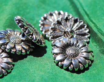 May include: Close-up of several silver-colored daisy-shaped buttons. The buttons are arranged on a vibrant green fabric. Each button features detailed petal and center designs, suggesting a vintage or handcrafted style.