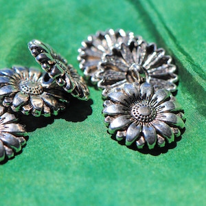 May include: Close-up of several silver-colored daisy-shaped buttons. The buttons are arranged on a vibrant green fabric. Each button features detailed petal and center designs, suggesting a vintage or handcrafted style.