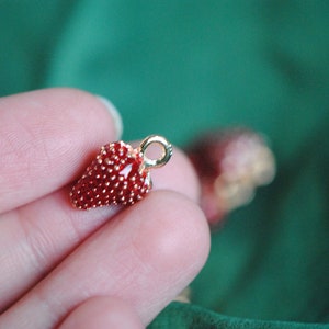 Pode incluir: Um amuleto dourado em forma de morango. O morango &eacute; vermelho e tem uma superf&iacute;cie texturizada. O amuleto tem um la&ccedil;o na parte superior para prender a um colar ou pulseira.