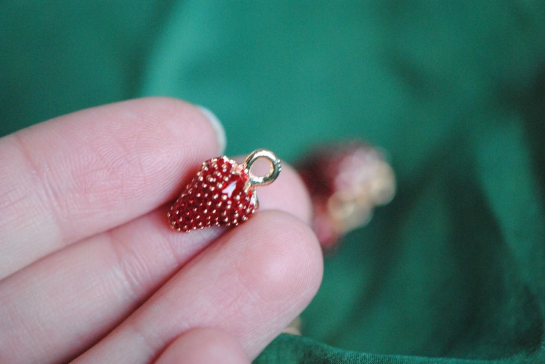 Pode incluir: Um amuleto dourado em forma de morango. O morango &eacute; vermelho e tem uma superf&iacute;cie texturizada. O amuleto tem um la&ccedil;o na parte superior para prender a um colar ou pulseira.