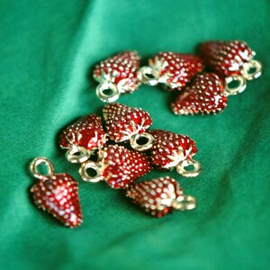 Pode incluir: Uma cole&ccedil;&atilde;o de 10 pingentes de morango vermelho com detalhes dourados. Os pingentes est&atilde;o dispostos sobre um fundo de tecido verde.