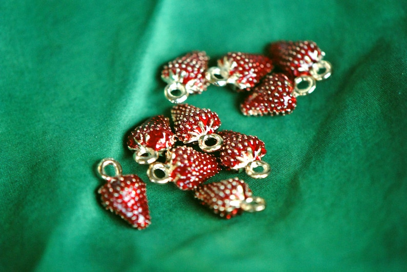 Pode incluir: Uma cole&ccedil;&atilde;o de 10 pingentes de morango vermelho com detalhes dourados. Os pingentes est&atilde;o dispostos sobre um fundo de tecido verde.