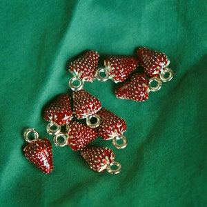 Pode incluir: Um grupo de pingentes de morango vermelho com la&ccedil;os de metal dourado. Os pingentes est&atilde;o espalhados sobre uma superf&iacute;cie de tecido verde.