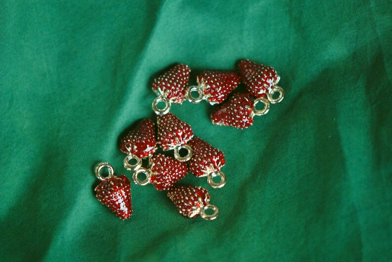 Pode incluir: Um grupo de pingentes de morango vermelho com la&ccedil;os de metal dourado. Os pingentes est&atilde;o espalhados sobre uma superf&iacute;cie de tecido verde.