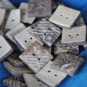 May include: A collection of square-shaped buttons made from natural coconut shell. The buttons have a light brown color with a darker brown pattern and two holes in the center.