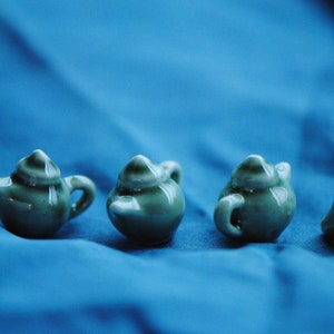 Pode incluir: Quatro pequenas chaleiras de cer&acirc;mica verde com acabamento esmaltado. As chaleiras est&atilde;o dispostas em fila sobre uma superf&iacute;cie azul.