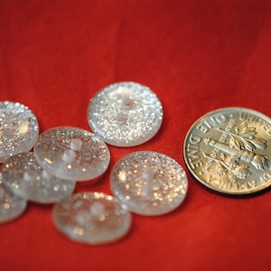 May include: A collection of eight clear glass buttons with a silver glitter finish. The buttons are arranged on a red surface with a silver dime for scale.