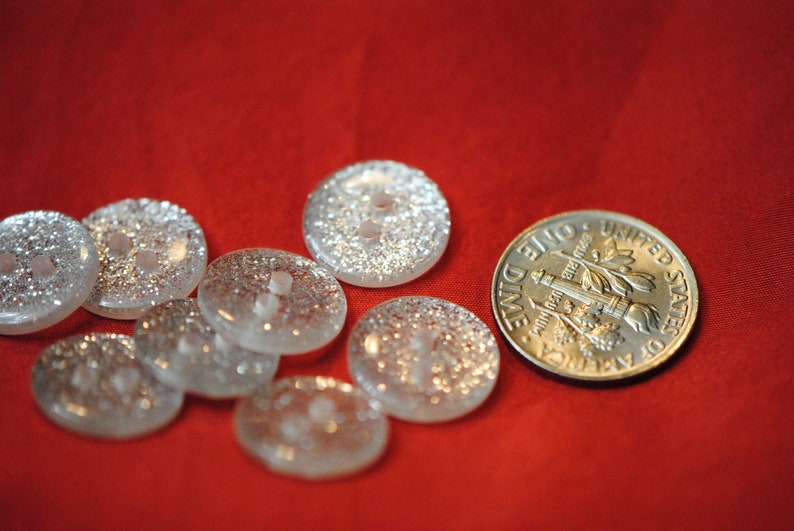 May include: A collection of eight clear glass buttons with a silver glitter finish. The buttons are arranged on a red surface with a silver dime for scale.