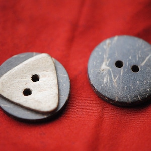 Op de afbeelding: Twee ronde, bruine kokosnootschelpknopen met elk twee gaten. Eén knop heeft een witte driehoek met twee gaten in het midden. De knopen liggen op een rode stoffen achtergrond.