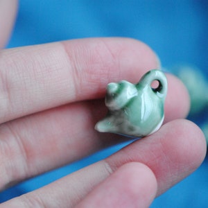Pode incluir: Um pequeno amuleto de chaleira de cer&acirc;mica verde com um esmalte branco. A chaleira tem uma al&ccedil;a e um bico. O amuleto est&aacute; sendo segurado em uma m&atilde;o.
