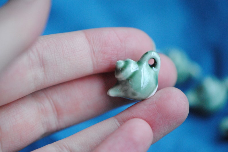 Pode incluir: Um pequeno amuleto de chaleira de cer&acirc;mica verde com um esmalte branco. A chaleira tem uma al&ccedil;a e um bico. O amuleto est&aacute; sendo segurado em uma m&atilde;o.