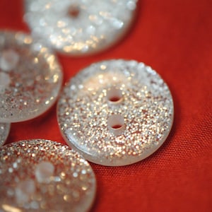 May include: A collection of six silver glitter buttons with two holes each. The buttons are arranged in a semi-circle on a red fabric background.