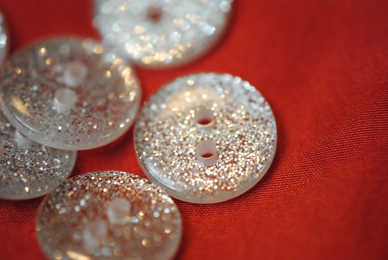 May include: A collection of six silver glitter buttons with two holes each. The buttons are arranged in a semi-circle on a red fabric background.