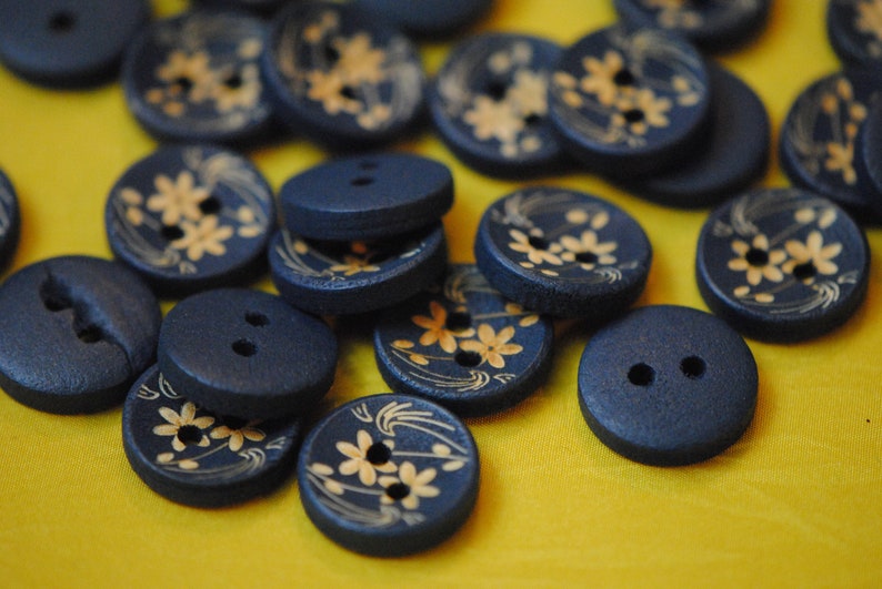 May include: A collection of dark blue buttons with a floral design in gold. The buttons are arranged on a yellow surface.