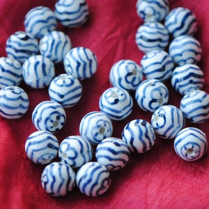 May include: A collection of spherical ceramic beads featuring a white base with hand-painted blue wavy lines and floral patterns. Each bead has a central hole for threading, showcasing a glossy finish and consistent round shape throughout the set.