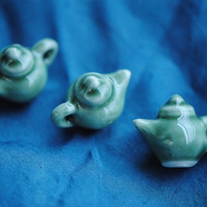 May include: Four small green ceramic teapots with different designs. The teapots are arranged on a blue fabric surface.