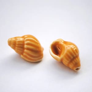 May include: Two small, ceramic seashells in shades of orange and tan sit on a white surface. The shells have detailed ridges and a glossy finish, suggesting a decorative or collectible item. The shells are oriented in different directions.