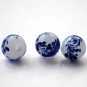 Puede incluir: Tres cuentas de vidrio blanco con diseños florales azules. Las cuentas son redondas y tienen un acabado brillante.
