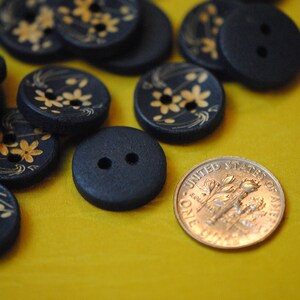 May include: A collection of black buttons with gold floral designs. Some buttons have two holes, while others have four holes. The buttons are arranged on a yellow surface.