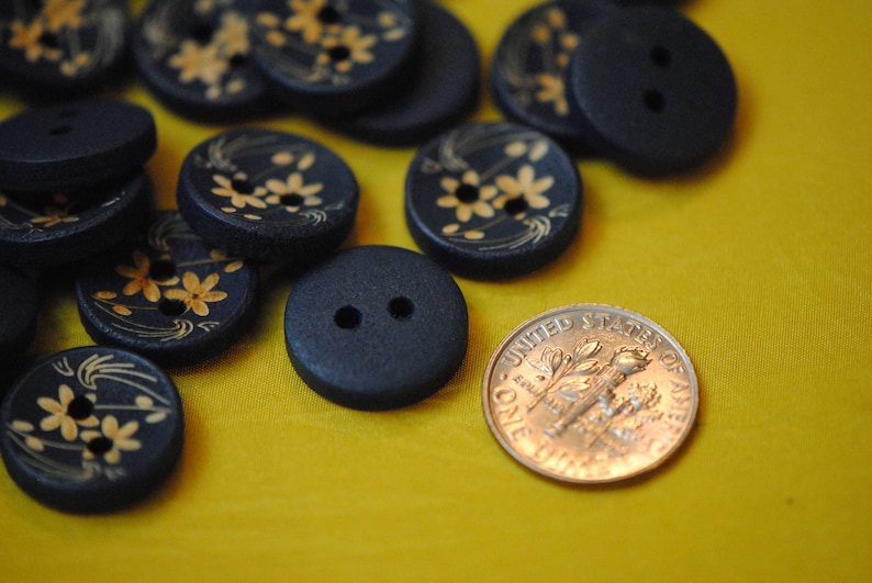 May include: A collection of black buttons with gold floral designs. Some buttons have two holes, while others have four holes. The buttons are arranged on a yellow surface.