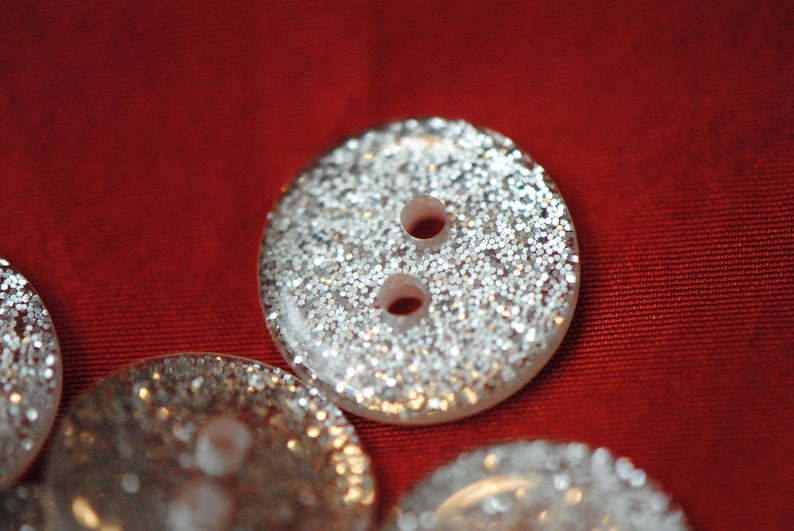 May include: Three silver glitter buttons with two holes each. The buttons are round and have a slightly raised edge.