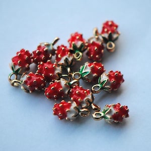 May include: A collection of small strawberry-shaped charms featuring bright red fruit with textured surfaces, white bases, and green leaf accents. Each charm includes a small gold-toned metal loop at the top for attachment, arranged in a cluster.