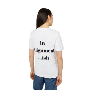 Op de afbeelding: Een wit T-shirt met korte mouwen en de zwarte tekst "In Alignment ...ish" op de achterkant. Het T-shirt wordt gecombineerd met een blauwe spijkerbroek. Het ontwerp is gecentreerd op de bovenrug van het kledingstuk in een vetgedrukt lettertype.