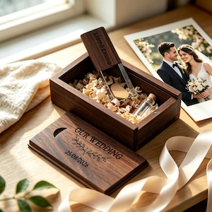 Peut inclure: Une boîte rectangulaire en bois foncé avec un couvercle coulissant gravé du texte OUR WEDDING, d'une branche de feuilles et de la date 25/08/2026. La boîte ouverte contient des copeaux de bois, une petite fiole en verre et une clé USB en bois.