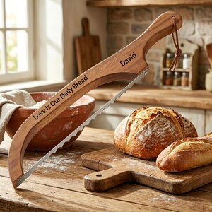 Könnte beinhalten: Ein Brotschneider aus Holz mit gezackter Metallklinge und einer Lederschlaufe am Griff. Das Holz ist mit dem Text "Love Is Our Daily Bread" und dem Namen "David" in einer dunklen, klaren Schriftart an der Seite des Griffs graviert.