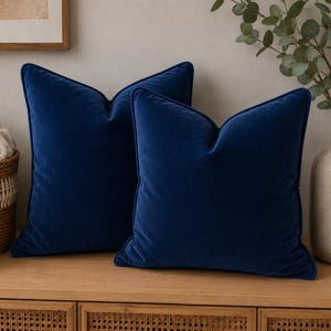 May include: Two square throw cushions in a deep navy blue velvet fabric. Each cushion features a soft, plush texture and a matching piped edge detail along the seams, creating a structured and uniform appearance for these decorative home accessories.