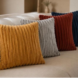 Pode incluir: Um conjunto de quatro almofadas quadradas dispostas em fila, cada uma com uma textura canelada vertical. As almofadas são de cor amarelo mostarda, cinzento claro, laranja queimado e azul marinho escuro, feitas de um tecido macio e felpudo.