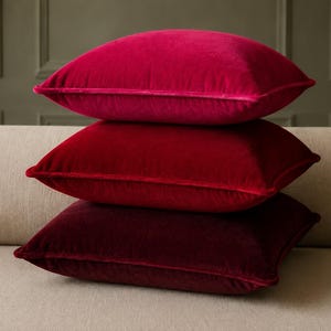 May include: A stack of three square velvet throw pillows in varying shades of red. The top pillow is a bright fuchsia, the middle pillow is a vibrant cherry red, and the bottom pillow is a deep, dark burgundy. Each pillow features piped edges.