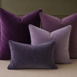 Puede incluir: Una colección de cuatro cojines de terciopelo cuadrados y rectangulares en varios tonos de morado, que van desde el violeta real profundo hasta un tono lavanda más claro, cada uno con un borde ribeteado y una textura suave.