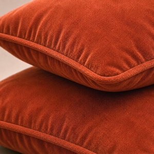 May include: Two square throw pillows stacked on top of each other, both featuring a solid burnt orange velvet fabric with a matching piped edge trim along the seams. The soft, plush texture of the velvet material is clearly visible on the surface.