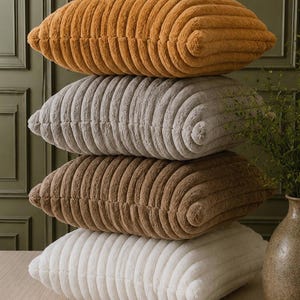May include: A stack of four square throw pillows featuring a textured, ribbed, plush surface. The pillows are arranged in a vertical column, displaying colors from top to bottom: mustard yellow, light gray, medium brown, and off-white.