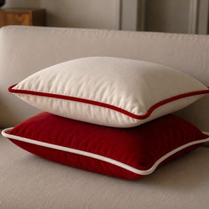 May include: Two square throw pillows stacked on top of each other. The top pillow is off-white with a vibrant red piped edge. The bottom pillow is deep red with a crisp white piped edge. Both pillows feature a soft, smooth fabric texture.