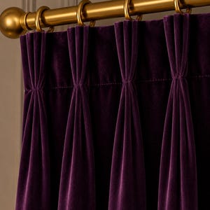 Op de afbeelding: Diep paars fluwelen gordijn met een ingeweven plooikop, opgehangen aan een gordijnroede van geborsteld goudkleurig metaal met bijpassende ronde ringen. De stof heeft een zachte, dichte textuur met verticale plooien die naar beneden vallen.