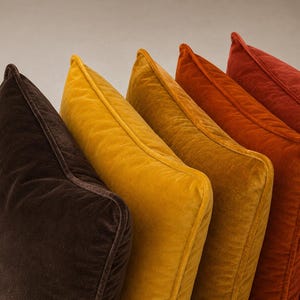 Può includere: Una fila di cinque cuscini quadrati in velluto disposti in una sfumatura di colori caldi. Le tonalità variano dal marrone scuro al giallo senape, giallo oro, arancione bruciato e rosso intenso. Ogni cuscino presenta bordi con profilo.