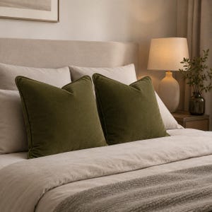 Op de afbeelding: Twee vierkante sierkussens in een diepe olijfgroene kleur met een zachte, textuurrijke stoffen afwerking. Deze kussens staan rechtop tegen een neutraal gekleurd hoofdeinde, rustend op een bed met lichtbeige beddengoed en een plaid.