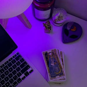 May include: A laptop, tarot cards, and decorative items are arranged on a white surface, illuminated by a purple light. The tarot cards feature illustrations of figures in ornate clothing. Other items include a candle, a small gold statue, and a purple alien-shaped dish.